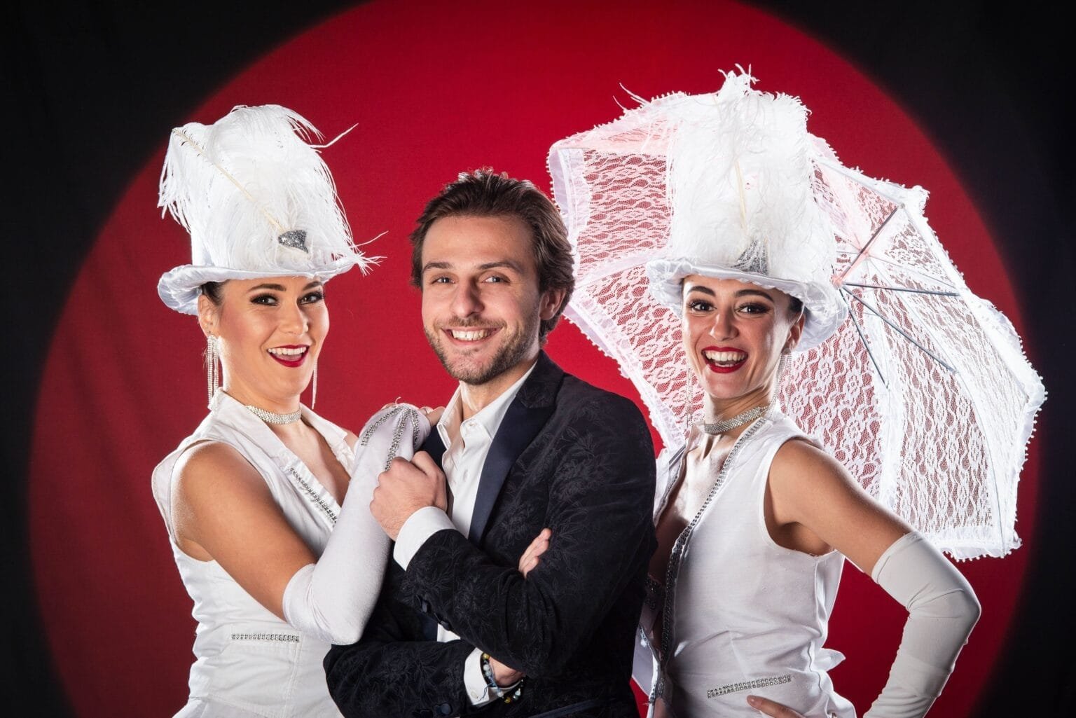 Le chanteur Benjamin Michel qui sourie face à la camera entourés de ces deux danseuse en costume blanc de scene et un parapluie en dentelle blanc pour accessoire