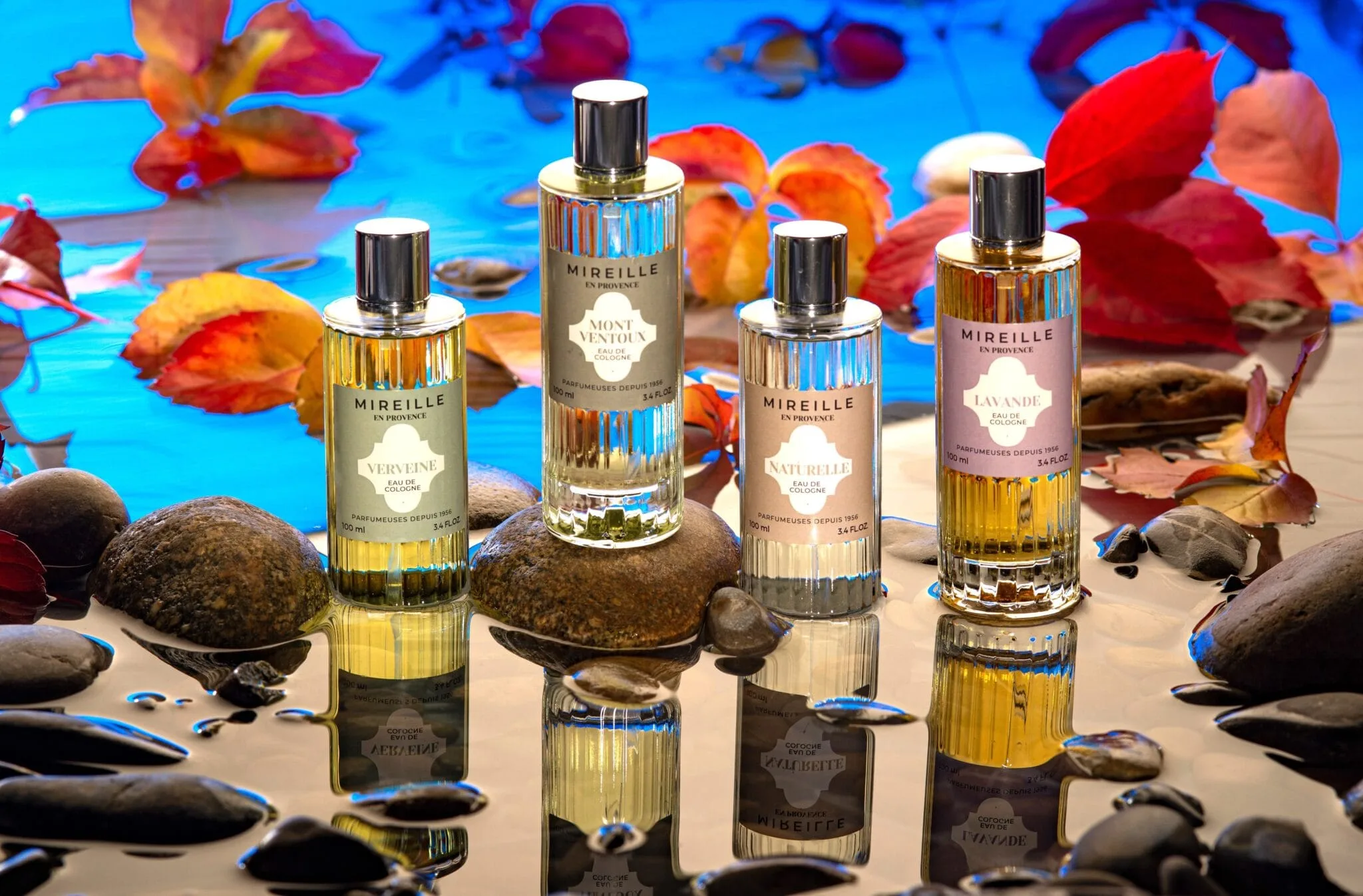 Quatre flacons de parfum avec leurs reflets sur l'eau entourées de galets de riviere et des feuillets aux couleurs d'automne